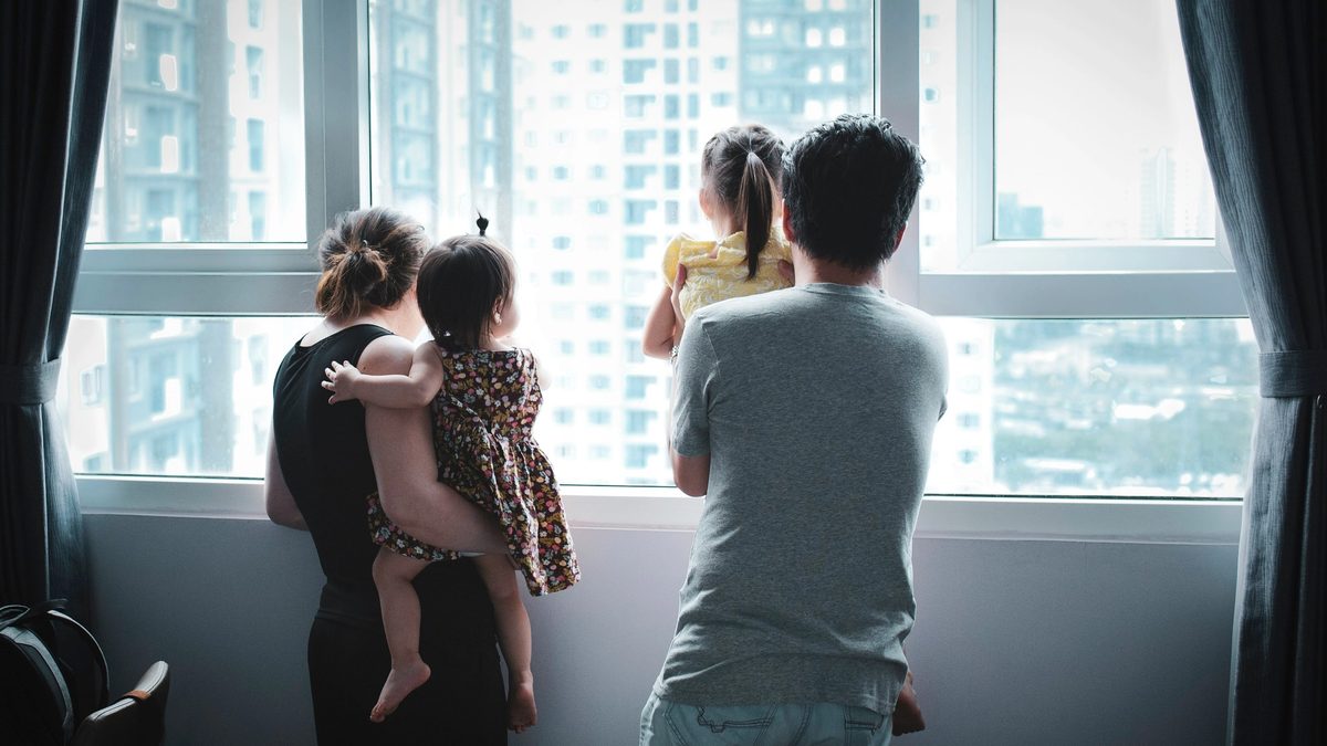 Family with two young children standing by a window and looking outside together.