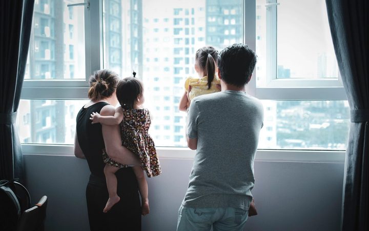 Family with two young children standing by a window and looking outside together.
