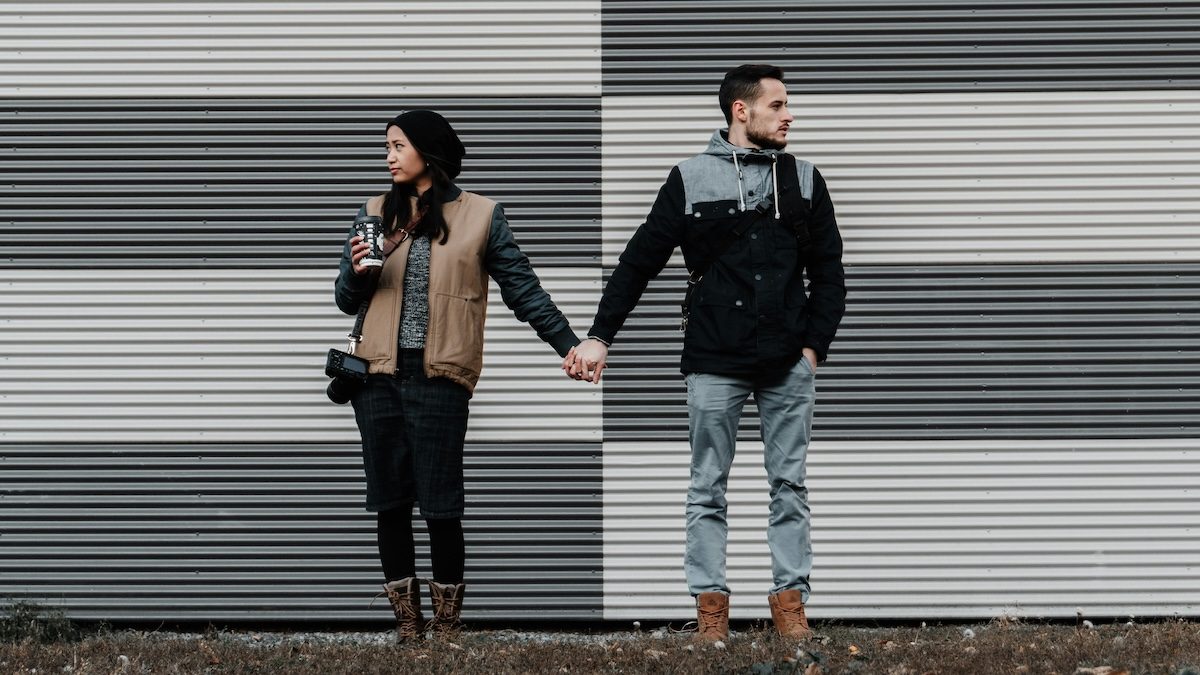 A couple where one person is avoidant and the other is anxious stands holding hands in front of a white and gray background for an article on attachment style and communicatio