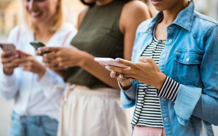 Three people standing outdoors, each looking down and using a smartphone.