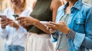 Three people standing outdoors, each looking down and using a smartphone.