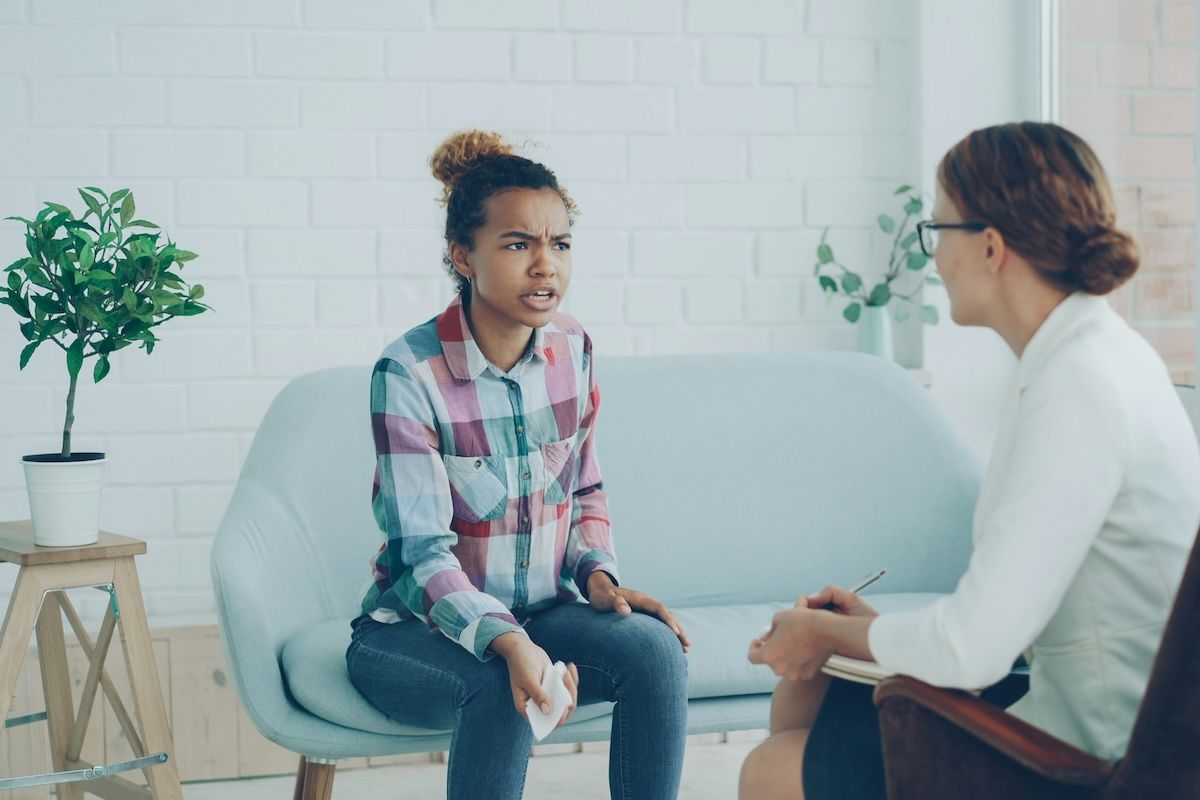 an angry teenage client is sitting on a therapy couch across from her therapist