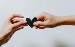 Two hands holding a black paper heart together.