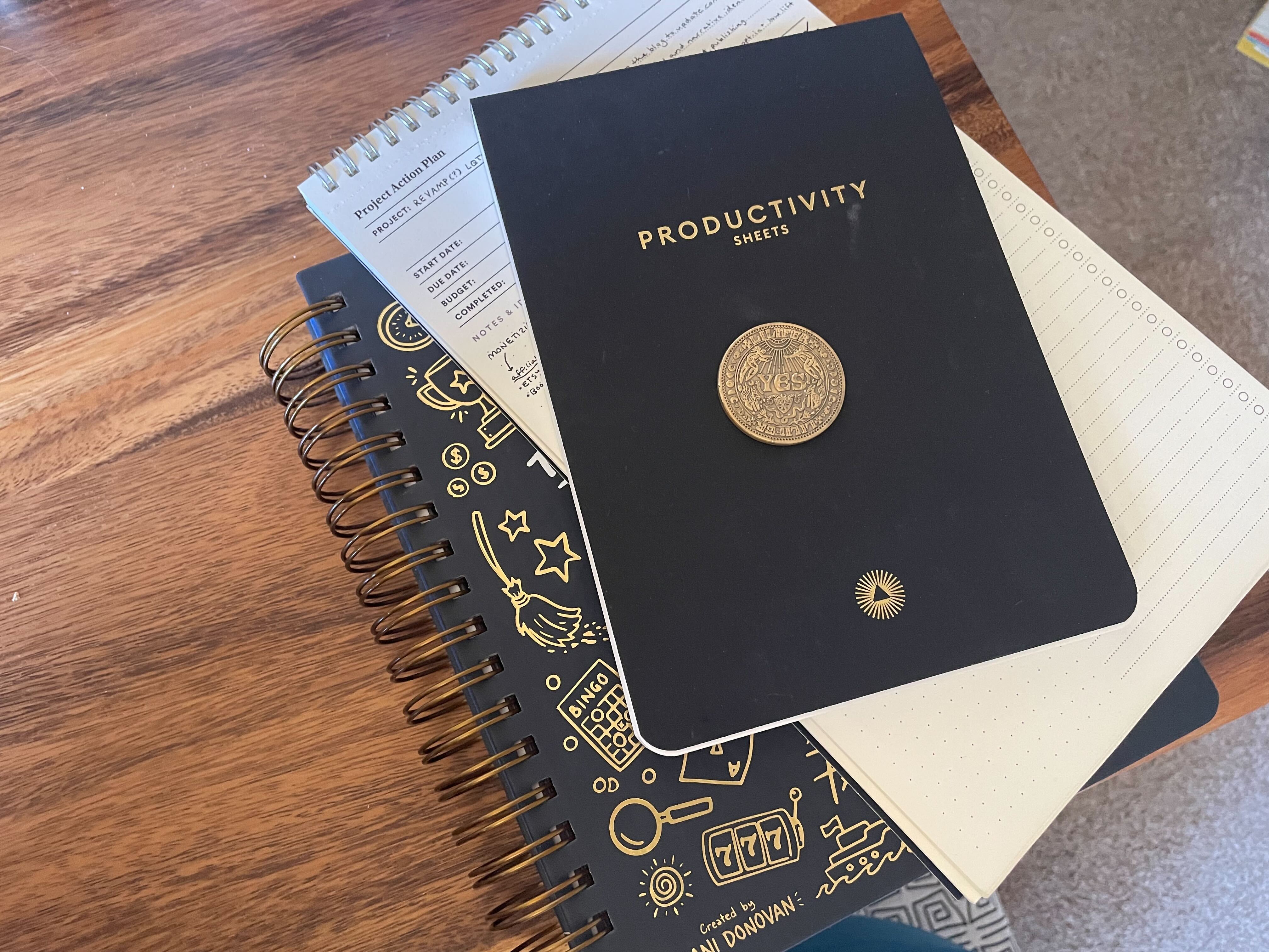 A stack of items on a desk, including a golden coin, two notepads, and a copy of Dani Donovan's "Anti-Planner" book peaking out from underneath.