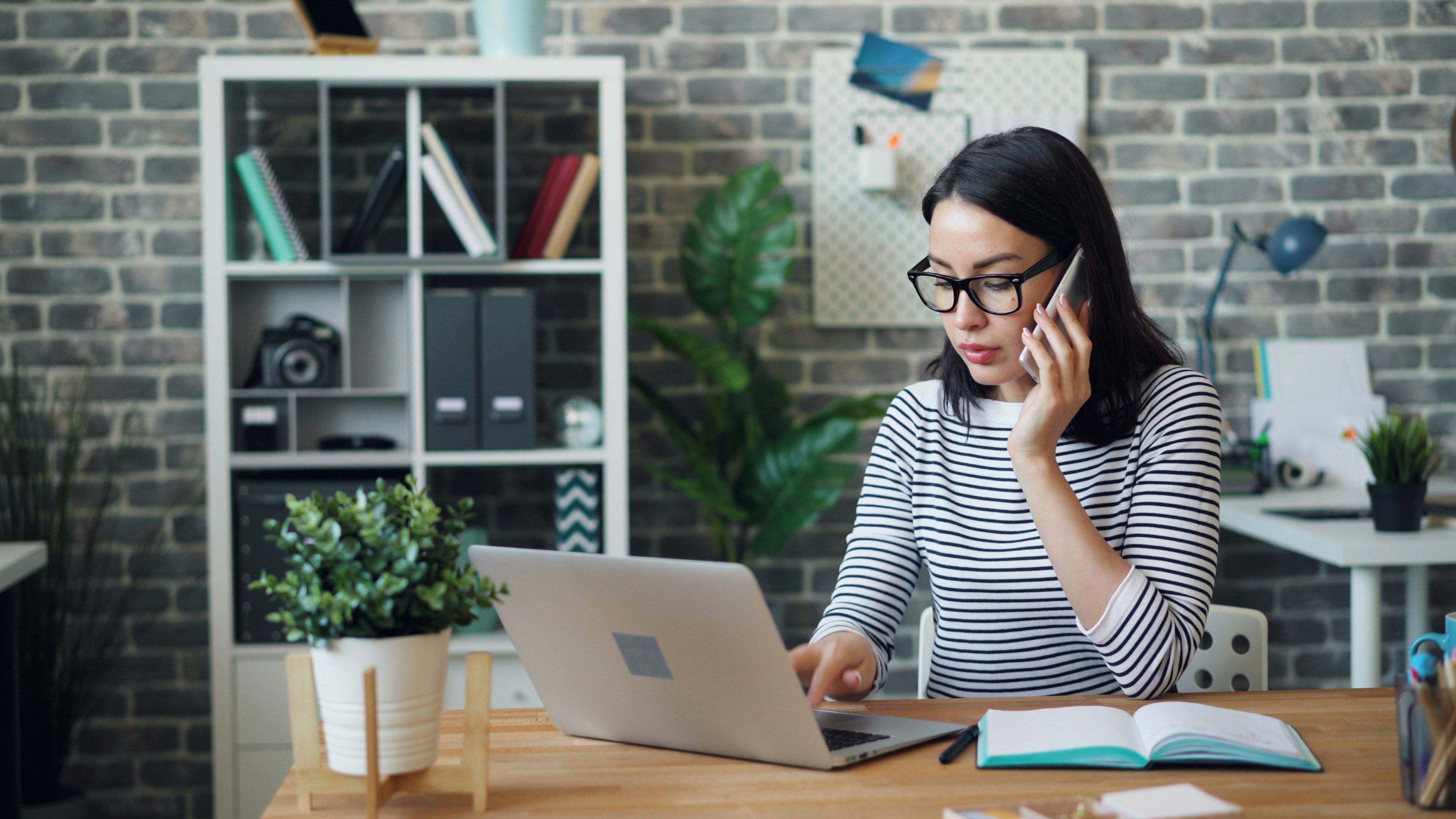Woman on a cellphone while working on a laptop at her desk.
