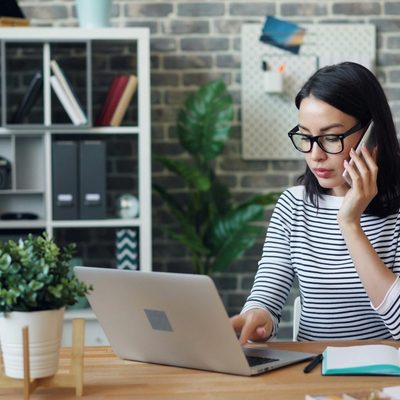 Woman on a cellphone while working on a laptop at her desk.