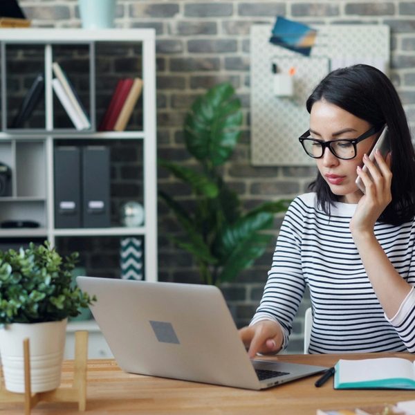 Woman on a cellphone while working on a laptop at her desk.