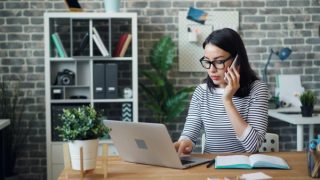 Woman on a cellphone while working on a laptop at her desk.