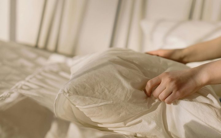 An image of two hands gently placing a pillow in white linens on a sun drenched bed.