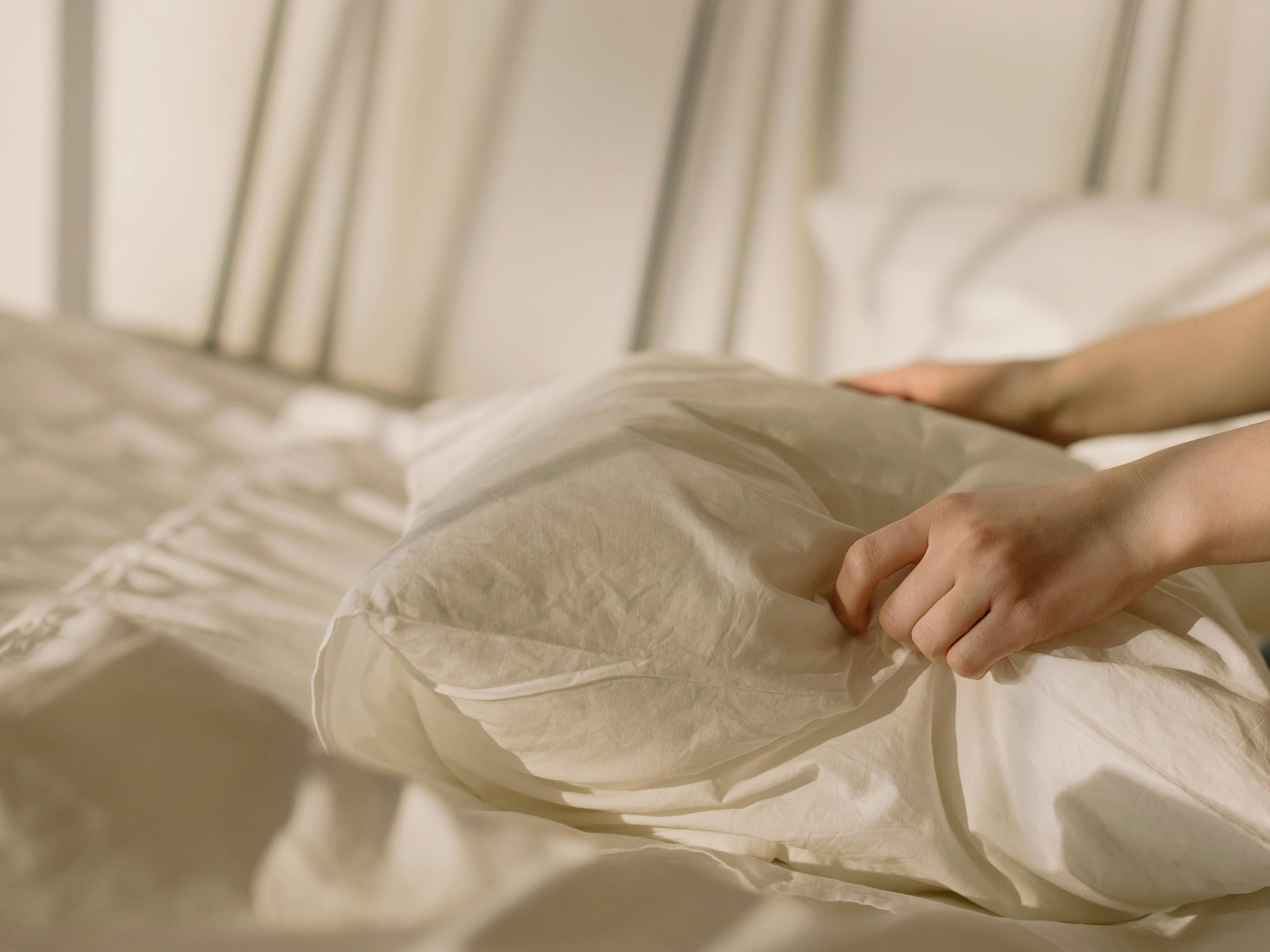 An image of two hands gently placing a pillow in white linens on a sun drenched bed.