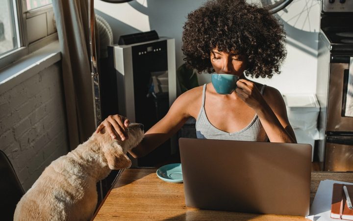 An Afro-Caribbean person finds calm before a virtual therapy session by sipping tea and petting their small, tawny dog.