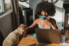An Afro-Caribbean person finds calm before a virtual therapy session by sipping tea and petting their small, tawny dog.