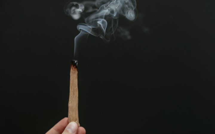 A hand holds a charred, smoking piece of palo santo wood in total darkness.