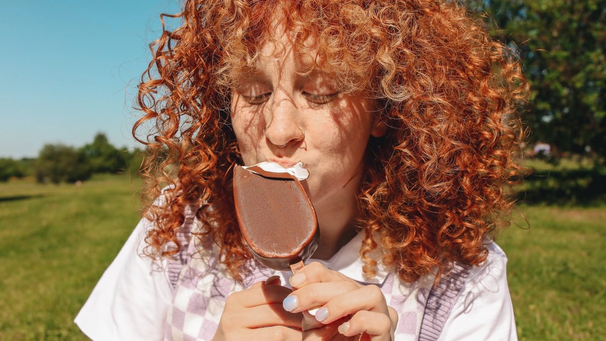 Sitting on a green, grassy lawn under a blue sky, a redhead enjoys an ice cream bar.