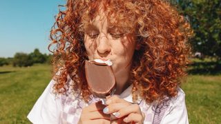 Sitting on a green, grassy lawn under a blue sky, a redhead enjoys an ice cream bar.