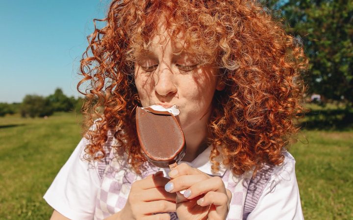 Sitting on a green, grassy lawn under a blue sky, a redhead enjoys an ice cream bar.