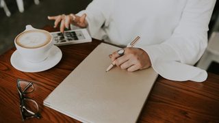 Person seated at a table holding pen on the left hand and browsing phone at the right, with a coffee cup and glasses on the table.