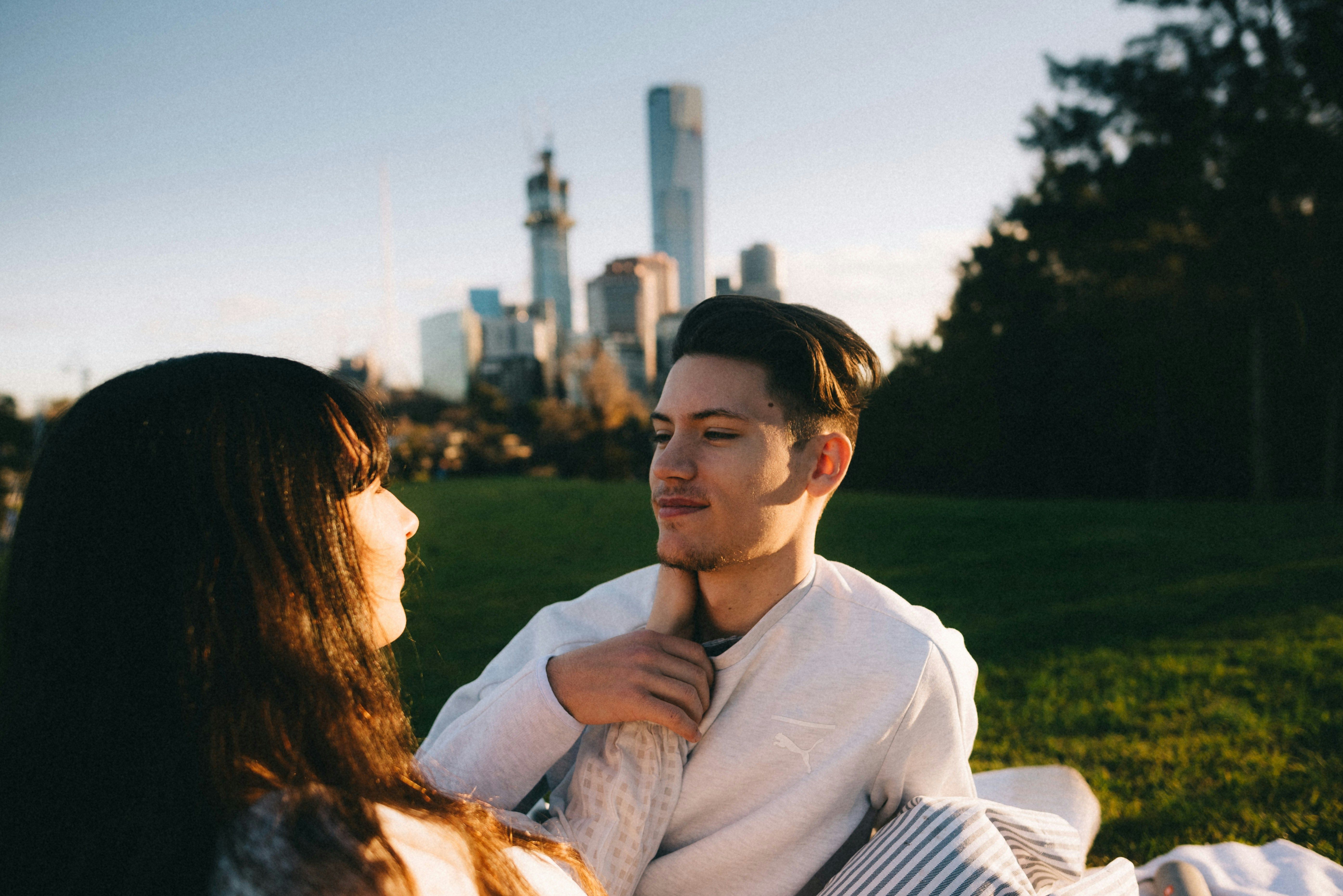 A couple sits outside in a grassy field, with tall city buildings fading on the horizon, as they stare into each other's eyes. One of them, with long dark hair, reaches to place their hand on the other's neck. The person with short hair smiles contentedly in return, placing a hand on the extended arm.