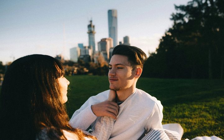 A couple sits outside in a grassy field, with tall city buildings fading on the horizon, as they stare into each other's eyes. One of them, with long dark hair, reaches to place their hand on the other's neck. The person with short hair smiles contentedly in return, placing a hand on the extended arm.