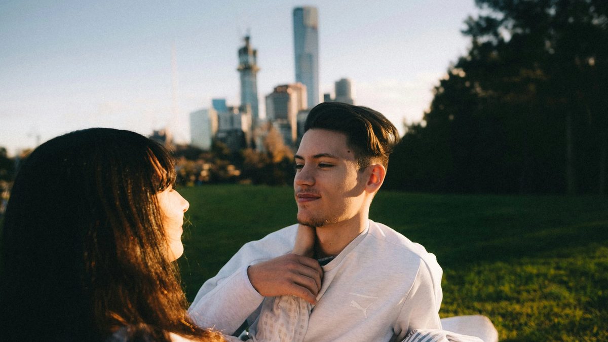 A couple sits outside in a grassy field, with tall city buildings fading on the horizon, as they stare into each other's eyes. One of them, with long dark hair, reaches to place their hand on the other's neck. The person with short hair smiles contentedly in return, placing a hand on the extended arm.