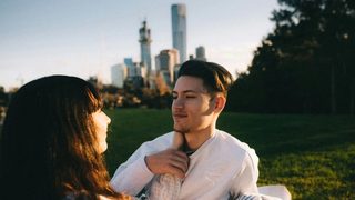 A couple sits outside in a grassy field, with tall city buildings fading on the horizon, as they stare into each other's eyes. One of them, with long dark hair, reaches to place their hand on the other's neck. The person with short hair smiles contentedly in return, placing a hand on the extended arm.