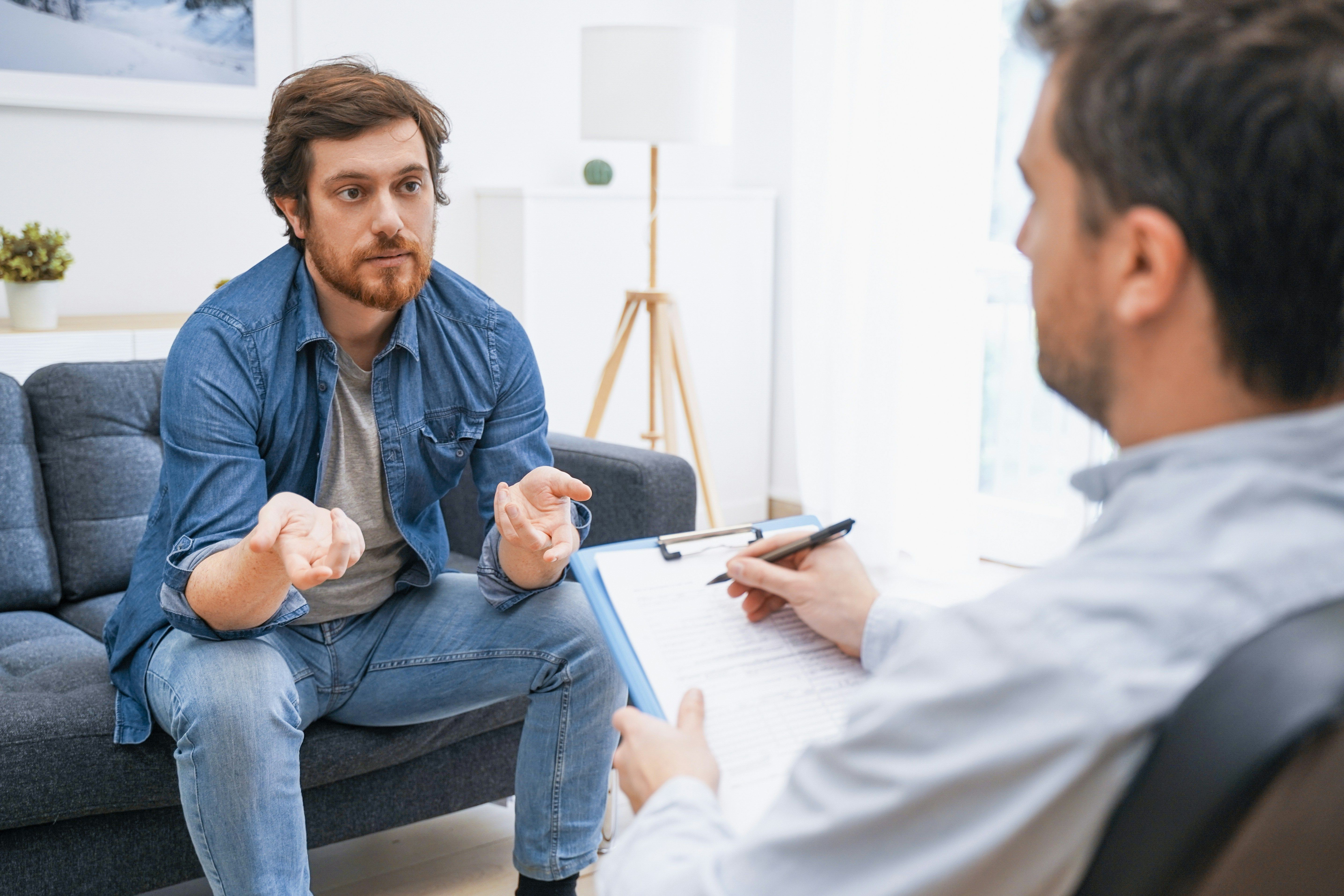 Therapist writing notes while male patient sits on a couch talking