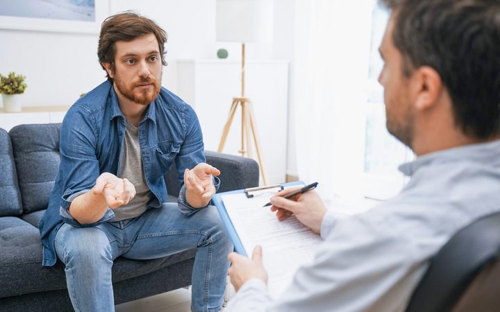 Therapist writing notes while male patient sits on a couch talking