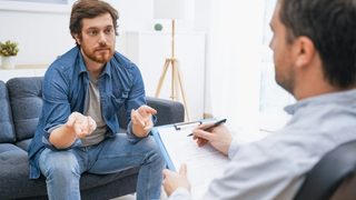 Therapist writing notes while male patient sits on a couch talking