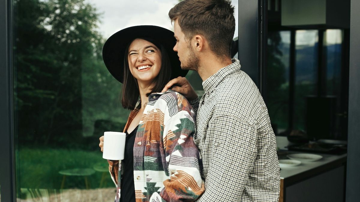 A young man and a woman stand together outside on the deck of a house in the countryside. The woman is smiling and looking inquisitively at the man.