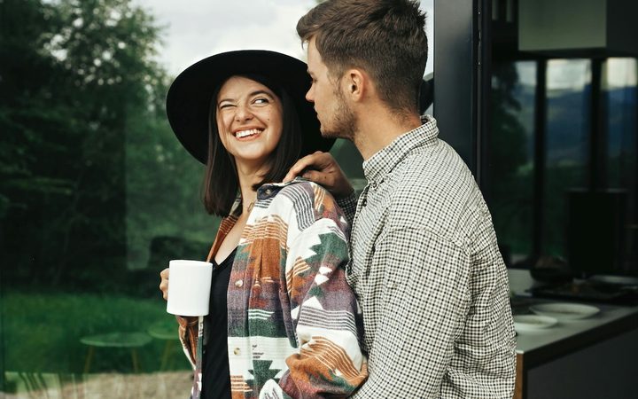 A young man and a woman stand together outside on the deck of a house in the countryside. The woman is smiling and looking inquisitively at the man.