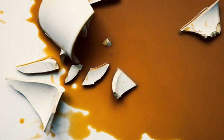 A broken ivory coffee mug on top of a puddle of spilled coffee on a white background