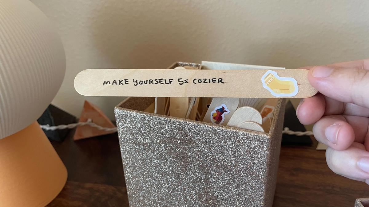A sparkly gold box filled with popsicle sticks. A hand retrieves a popsicle stick that has writing on it: "Make yourself 5% cozier." It has a sticker next to the writing, featuring a pair of yellow socks.
