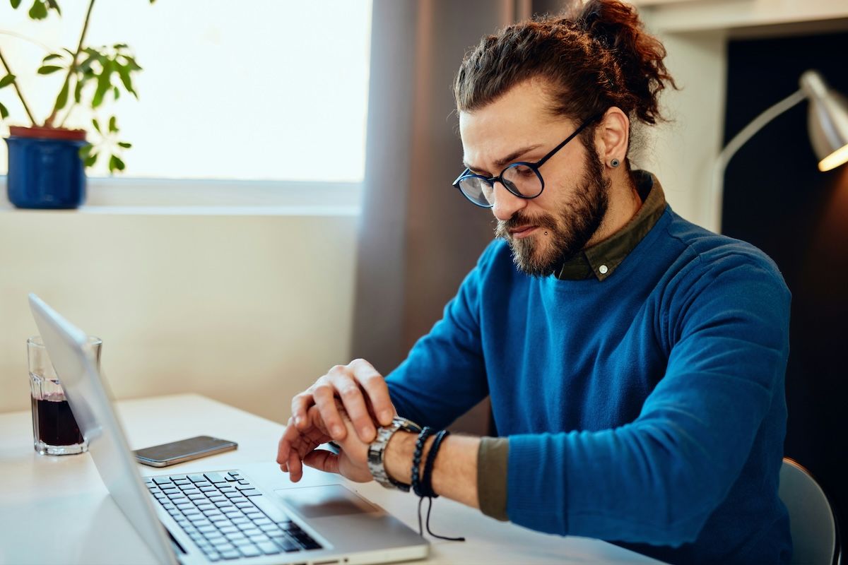 a therapist in a blue sweater is sitting at their desk, checking their watch after an online session