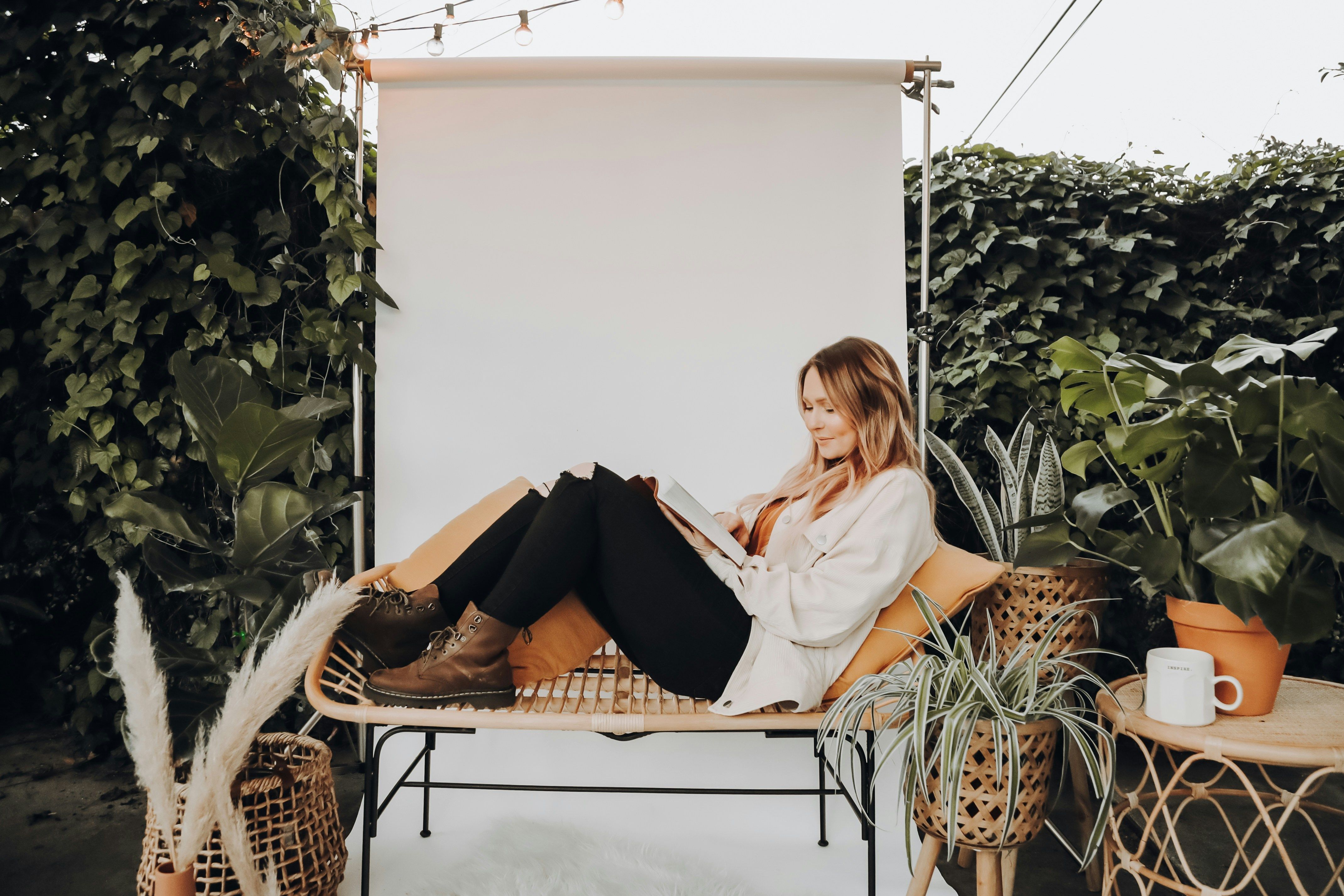 Woman reading on a rattan bench surrounded by plants and string lights in a cozy outdoor setting.
