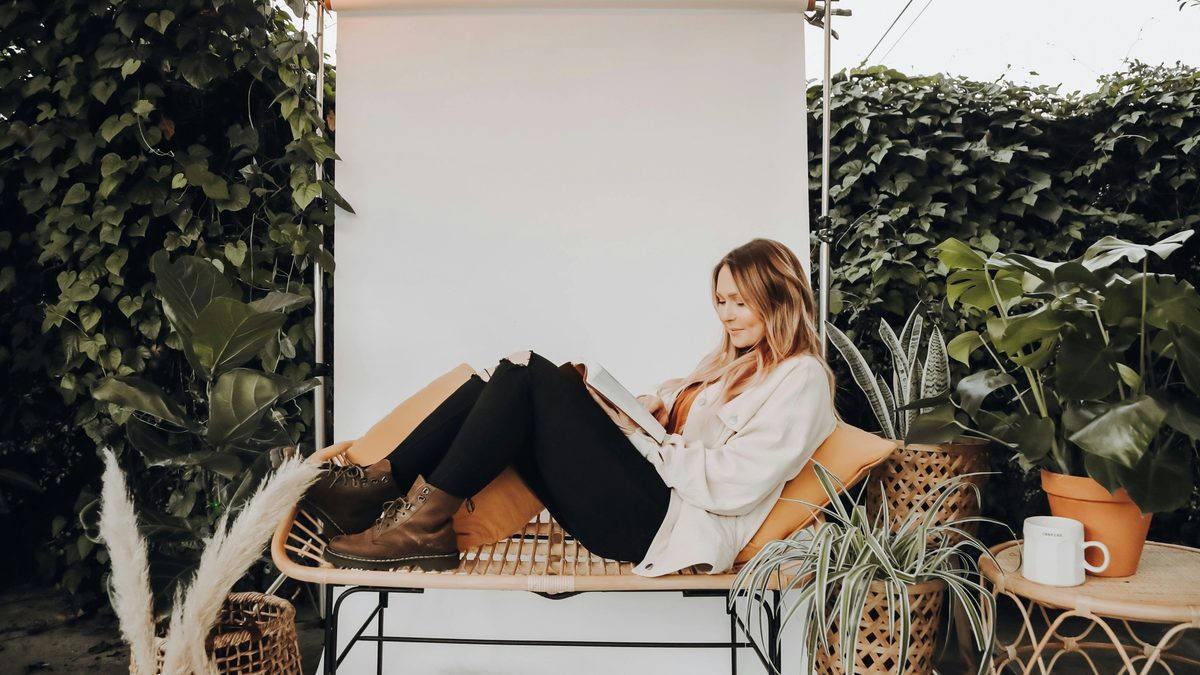 Woman reading on a rattan bench surrounded by plants and string lights in a cozy outdoor setting.