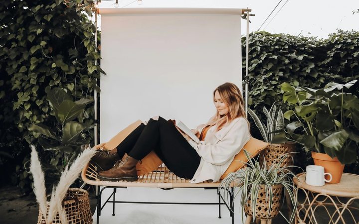 Woman reading on a rattan bench surrounded by plants and string lights in a cozy outdoor setting.