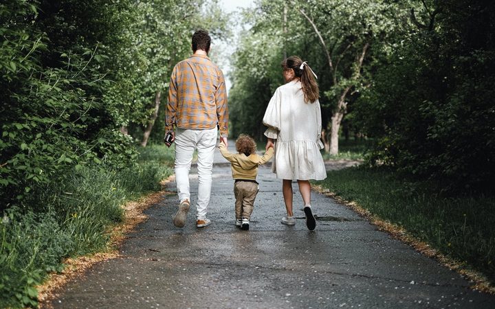 Two parents walk away from the camera along a paved path in the woods. In between them is their toddler, clasping each of their hands.