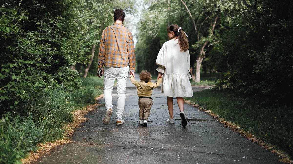 Two parents walk away from the camera along a paved path in the woods. In between them is their toddler, clasping each of their hands.