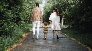 Two parents walk away from the camera along a paved path in the woods. In between them is their toddler, clasping each of their hands.