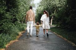 Two parents walk away from the camera along a paved path in the woods. In between them is their toddler, clasping each of their hands.