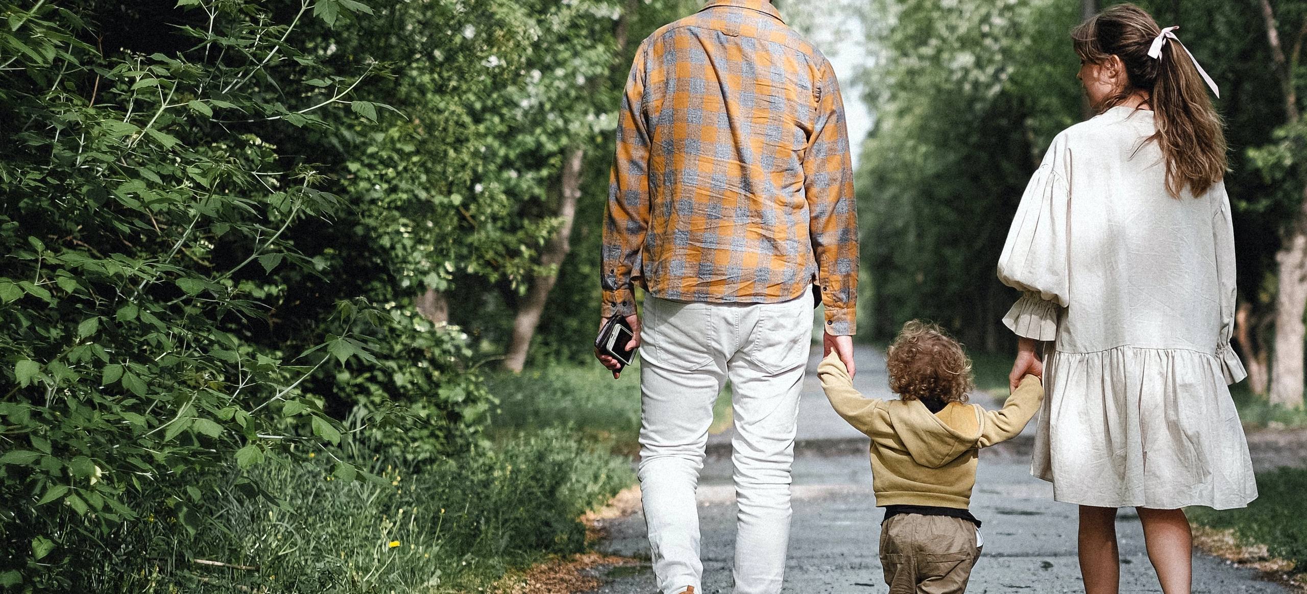 Two adults walking hand in hand with a toddler down a tree-lined path, seen from behind