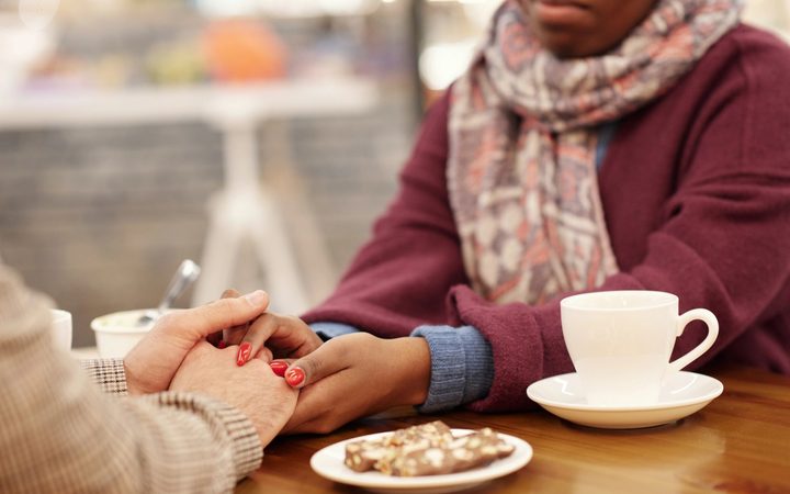 Over a table with a pastry and coffee, a woman holds her friends hand as a show of support.