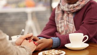 Over a table with a pastry and coffee, a woman holds her friends hand as a show of support.