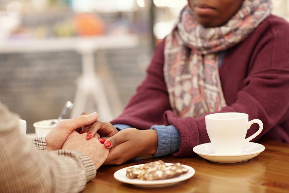 Over a table with a pastry and coffee, a woman holds her friends hand as a show of support.