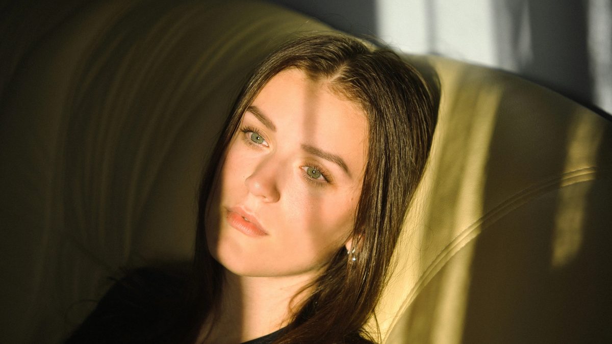 In a dark room after an online therapy appointment, a woman looks dejectedly through a sun-soaked window, throwing shadows across her face.