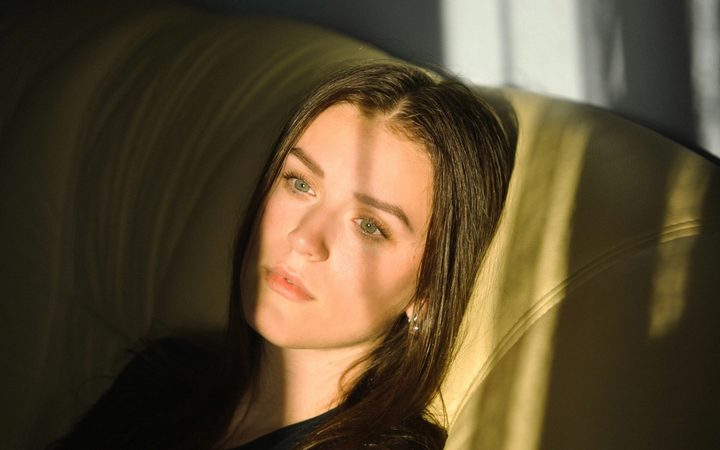 In a dark room after an online therapy appointment, a woman looks dejectedly through a sun-soaked window, throwing shadows across her face.