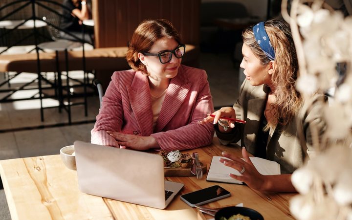 An experienced Latina therapist sits with a young mentee at a sun-drenched table inside a quiet café.