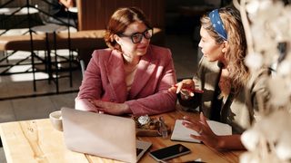 An experienced Latina therapist sits with a young mentee at a sun-drenched table inside a quiet café.