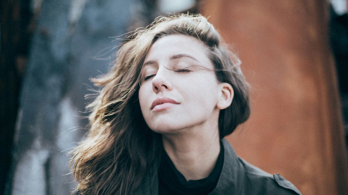 a calm, centered woman stands with her eyes closed outdoors, enjoying the late afternoon sun