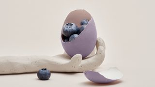 An outstretched hand made of clay, holding a purple egg whose shell has been cracked to reveal "superfood" blueberries inside.